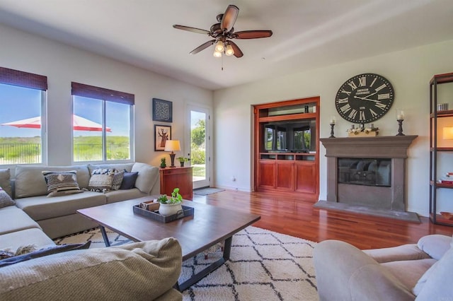 living area featuring a glass covered fireplace, wood finished floors, baseboards, and ceiling fan
