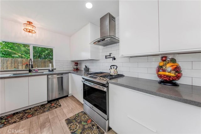 kitchen with wall chimney range hood, backsplash, appliances with stainless steel finishes, a sink, and white cabinets