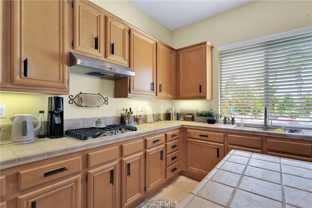 kitchen with a sink, tile counters, stainless steel gas stovetop, and under cabinet range hood