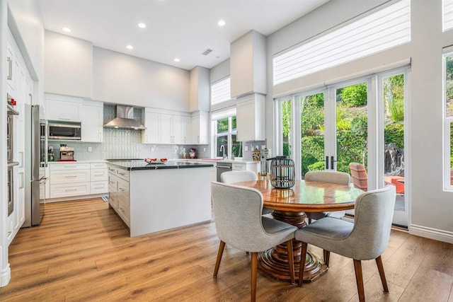kitchen featuring a towering ceiling, a center island, wall chimney exhaust hood, white cabinetry, and stainless steel appliances