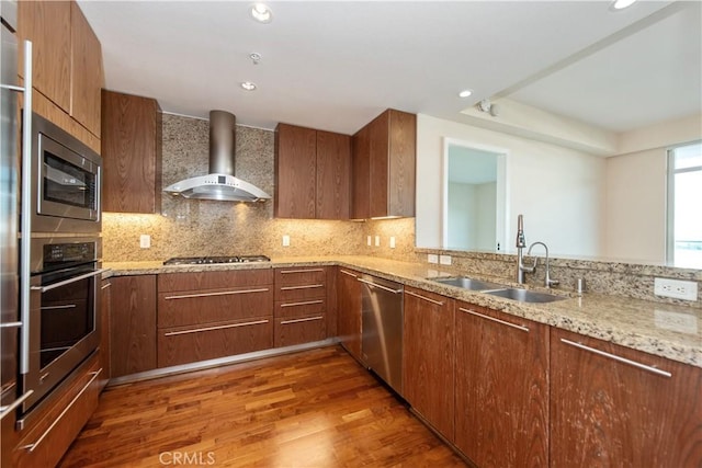 kitchen with wall chimney range hood, light stone counters, a sink, appliances with stainless steel finishes, and wood finished floors