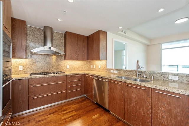 kitchen with wood finished floors, brown cabinetry, appliances with stainless steel finishes, wall chimney range hood, and a sink