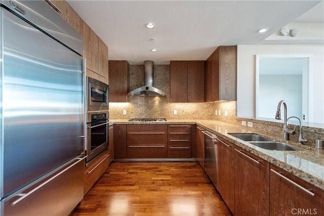 kitchen featuring a sink, built in appliances, light stone countertops, wall chimney exhaust hood, and wood finished floors