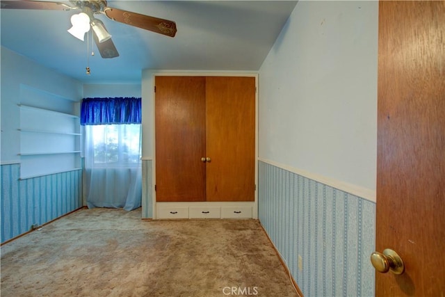 unfurnished bedroom featuring wallpapered walls, a wainscoted wall, carpet flooring, and a ceiling fan