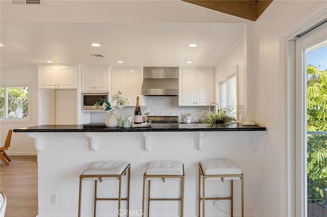 kitchen featuring a breakfast bar, visible vents, stainless steel microwave, wall chimney exhaust hood, and white cabinetry