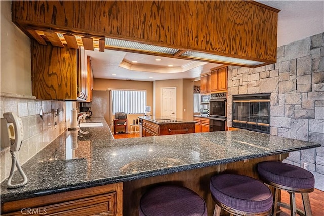 kitchen featuring dark stone countertops, black appliances, brown cabinets, a raised ceiling, and a sink