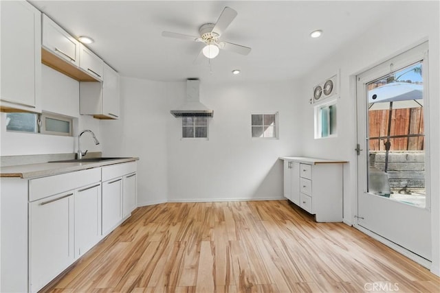 kitchen featuring light wood-style flooring, white cabinetry, wall chimney exhaust hood, ceiling fan, and a sink