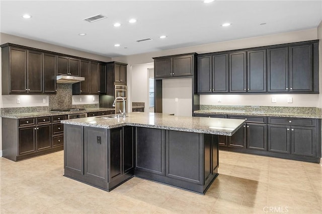 kitchen featuring light stone counters, under cabinet range hood, visible vents, a sink, and a center island with sink