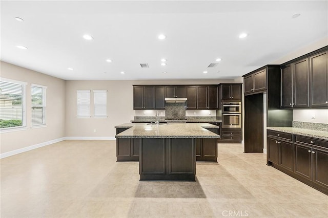 45600 Gresham, Temecula, CA 92592 - kitchen with a sink, a kitchen island with sink, light stone counters, dark brown cabinets, and baseboards