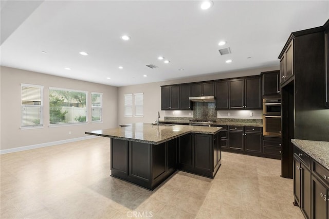 kitchen with an island with sink, under cabinet range hood, dark stone countertops, visible vents, and a sink