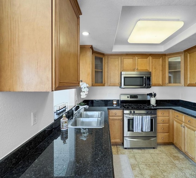 kitchen featuring brown cabinetry, a sink, a raised ceiling, glass insert cabinets, and stainless steel appliances