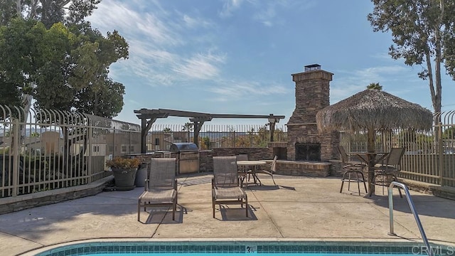 community pool with exterior kitchen, an outdoor stone fireplace, fence, a patio, and a grill