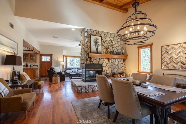 dining room with ceiling fan with notable chandelier, beamed ceiling, a stone fireplace, visible vents, and wood-type flooring
