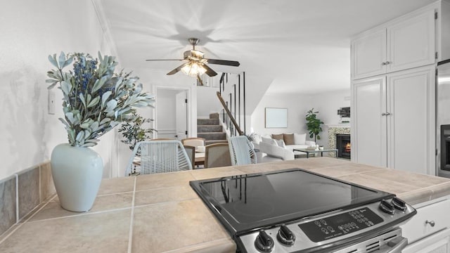 kitchen with ceiling fan, stainless steel range with electric cooktop, white cabinets, a warm lit fireplace, and tile counters