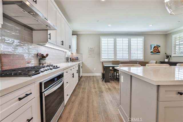 kitchen featuring exhaust hood, appliances with stainless steel finishes, ornamental molding, and light wood finished floors