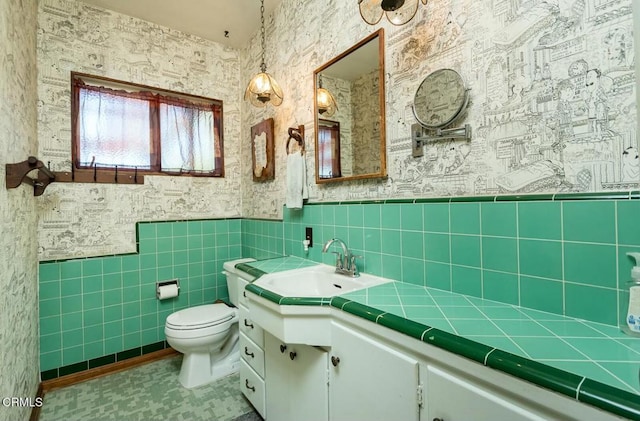 bathroom with vanity, wallpapered walls, toilet, wainscoting, and tile walls