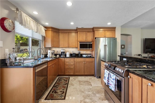 kitchen with arched walkways, a sink, brown cabinetry, recessed lighting, and stainless steel appliances
