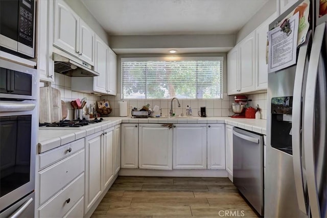 kitchen featuring a sink, under cabinet range hood, tile counters, white cabinetry, and stainless steel appliances