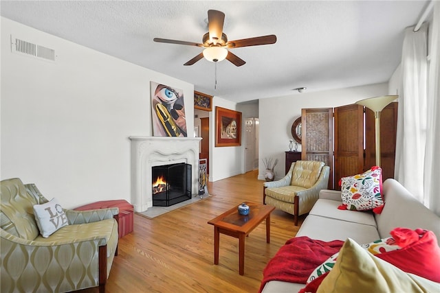 living area with light wood-type flooring, visible vents, ceiling fan, and a warm lit fireplace