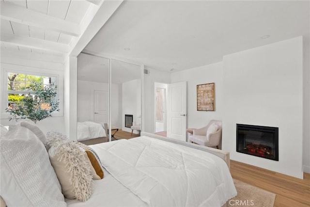 bedroom featuring a glass covered fireplace, light wood-style floors, and beam ceiling
