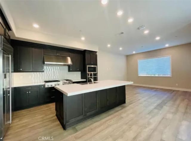 kitchen featuring light wood-style flooring, under cabinet range hood, a sink, a center island with sink, and built in appliances
