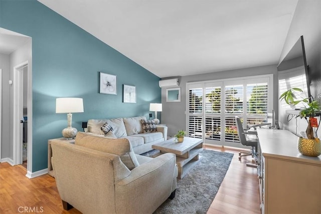 living area with an AC wall unit, baseboards, light wood finished floors, and vaulted ceiling