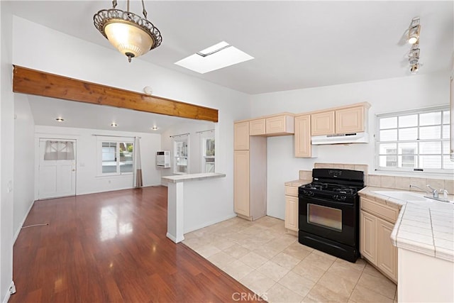 kitchen with a sink, black gas stove, vaulted ceiling with skylight, tile counters, and under cabinet range hood