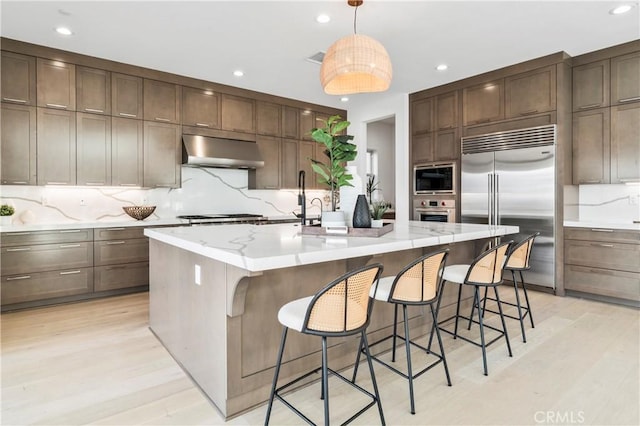 kitchen featuring an island with sink, light wood-type flooring, ventilation hood, decorative backsplash, and built in appliances