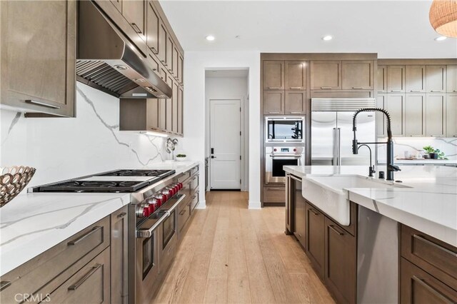 kitchen with built in appliances, light stone countertops, under cabinet range hood, light wood-style floors, and a sink
