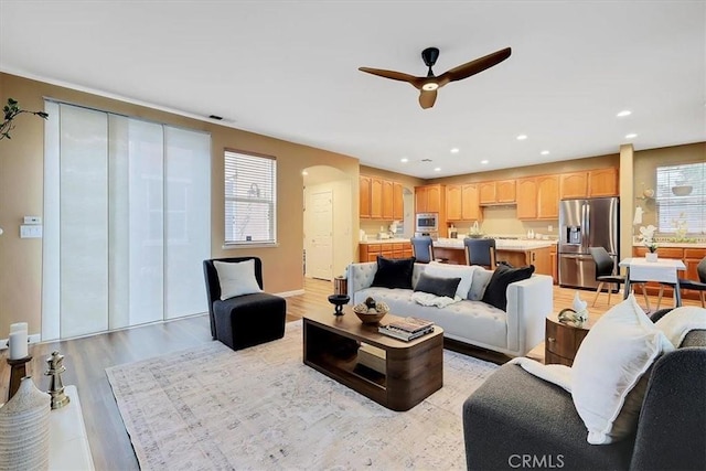 living area featuring arched walkways, light wood finished floors, a ceiling fan, and a healthy amount of sunlight