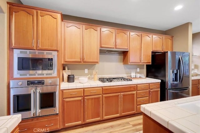 kitchen with light wood finished floors, tile counters, appliances with stainless steel finishes, and under cabinet range hood
