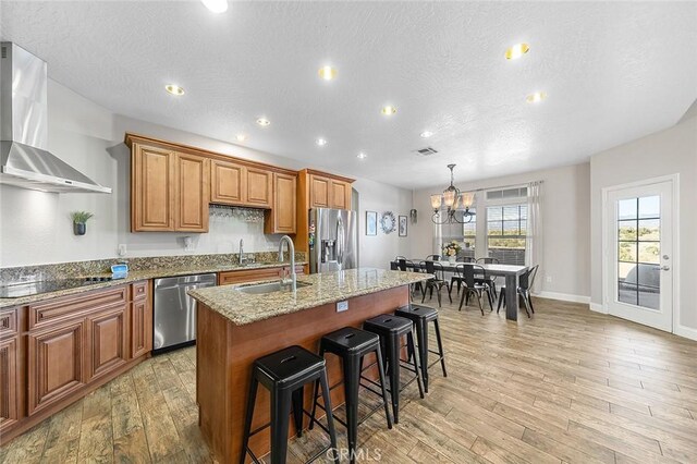 7922 Los Palmas Dr, Oak Hills, CA 92344 - kitchen with wall chimney exhaust hood, brown cabinetry, appliances with stainless steel finishes, a kitchen island with sink, and a sink
