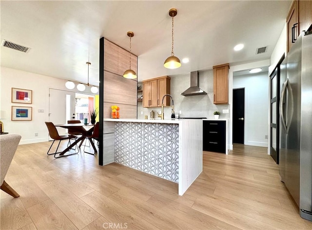 kitchen with light wood-style floors, freestanding refrigerator, a peninsula, wall chimney range hood, and visible vents
