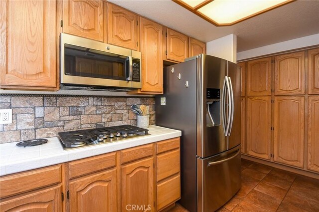 kitchen with backsplash, brown cabinets, dark tile patterned flooring, tile counters, and stainless steel appliances