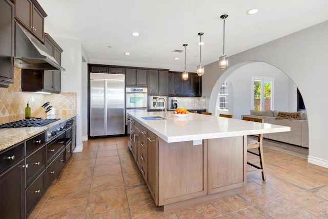 kitchen with arched walkways, a sink, light countertops, an island with sink, and stainless steel appliances