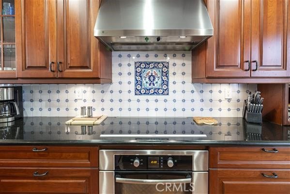 kitchen featuring brown cabinetry, decorative backsplash, wall chimney exhaust hood, and stainless steel oven
