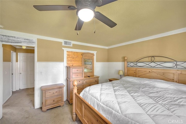bedroom featuring visible vents, ornamental molding, a ceiling fan, a wainscoted wall, and light colored carpet