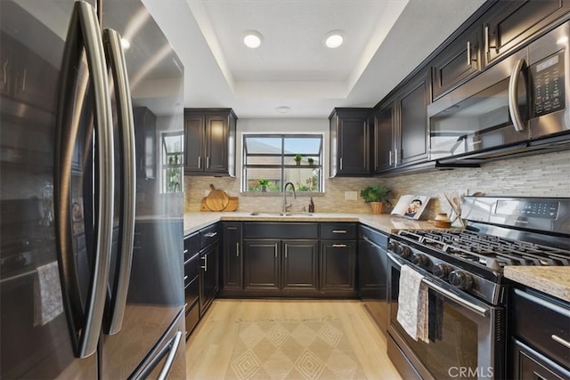 kitchen featuring a sink, a raised ceiling, tasteful backsplash, light wood-type flooring, and appliances with stainless steel finishes