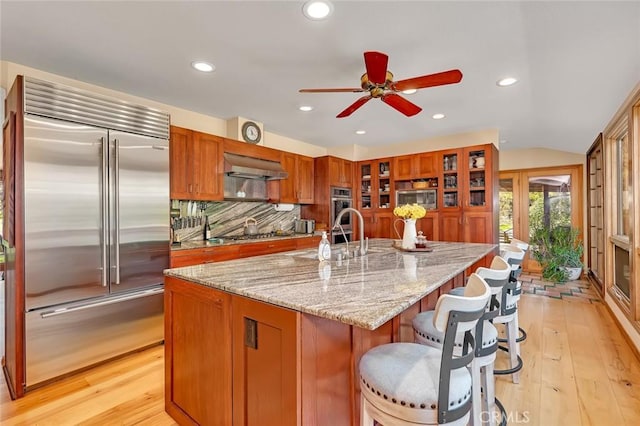 kitchen with built in appliances, ceiling fan, under cabinet range hood, brown cabinets, and light wood finished floors