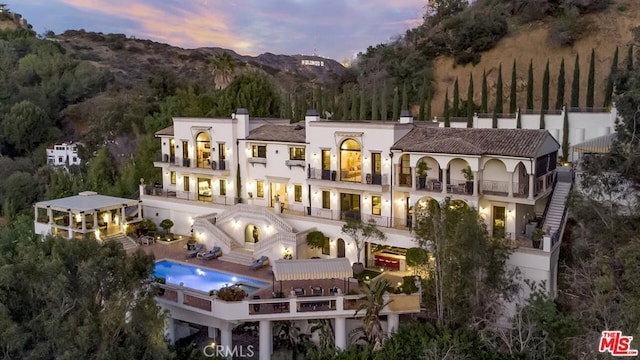 back of house at dusk with stucco siding, a patio, a balcony, and a chimney