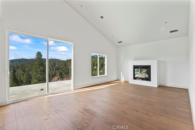 unfurnished living room featuring high vaulted ceiling, a view of trees, a glass covered fireplace, visible vents, and hardwood / wood-style floors