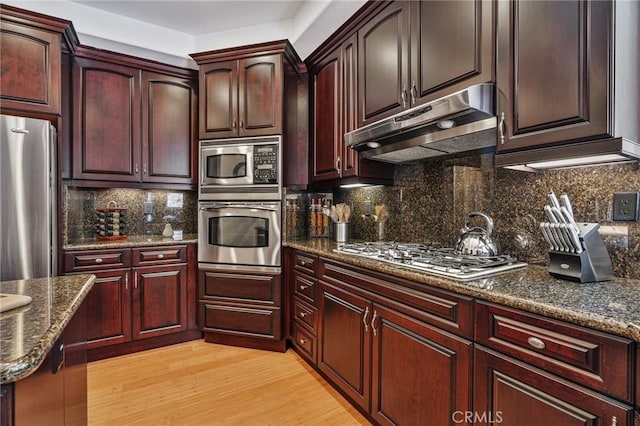 kitchen featuring appliances with stainless steel finishes, under cabinet range hood, tasteful backsplash, dark stone counters, and light wood-type flooring