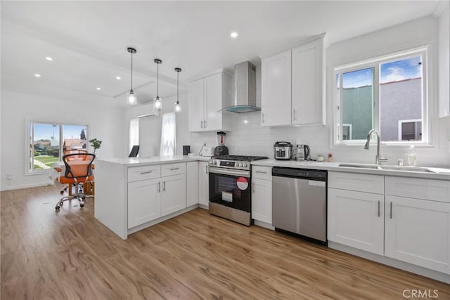 kitchen with a peninsula, light wood-style floors, wall chimney exhaust hood, a sink, and appliances with stainless steel finishes