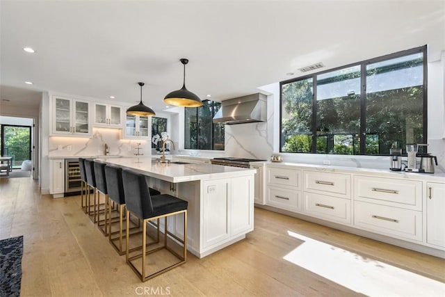 kitchen with light wood-style floors, wall chimney exhaust hood, glass insert cabinets, a sink, and beverage cooler