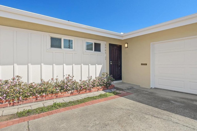 view of exterior entry with an attached garage, board and batten siding, driveway, and stucco siding