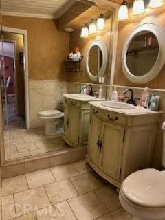 bathroom featuring vanity, ornamental molding, toilet, wainscoting, and tile walls