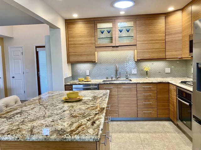 kitchen featuring light stone counters, a sink, brown cabinetry, stainless steel appliances, and backsplash