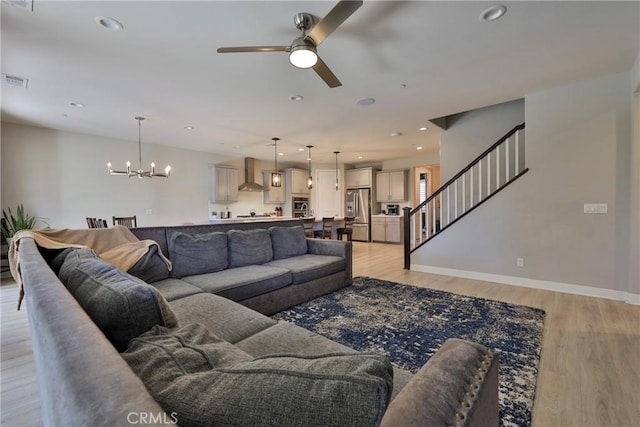 living area featuring light wood-type flooring, ceiling fan with notable chandelier, visible vents, and stairway