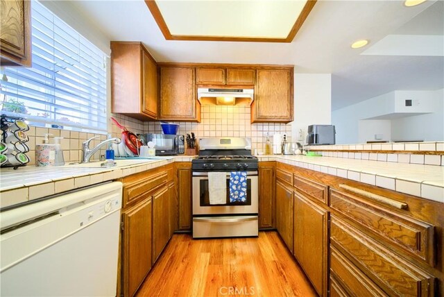 kitchen with white dishwasher, a sink, stainless steel gas range, under cabinet range hood, and brown cabinets