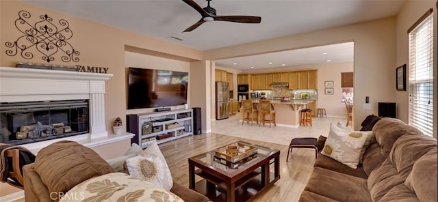 living area featuring light wood-style flooring, a glass covered fireplace, a ceiling fan, visible vents, and recessed lighting